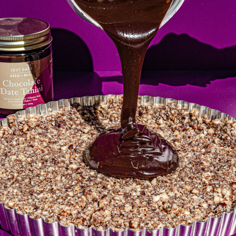 Chocolate ganache being poured from a glass measuring cup into a pecan tart crust, beside a jar of Just Date Chocolate Date Tahini