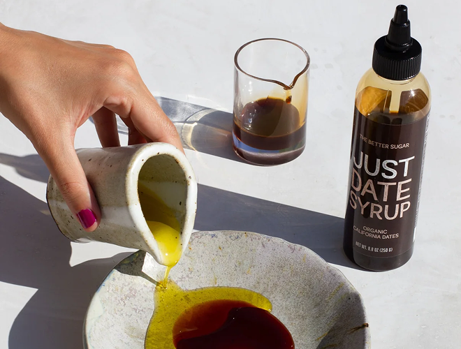 Person pouring syrup from a container into a dish with a bottle of 'Just Date Syrup' in the background.