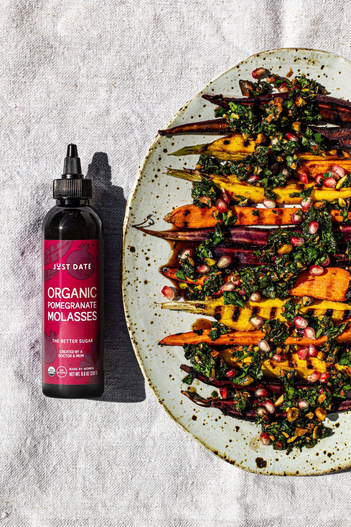Bottle of Just Date Organic Pomegranate Molasses next to a plate of roasted vegetables with pomegranate seeds on a textured surface.