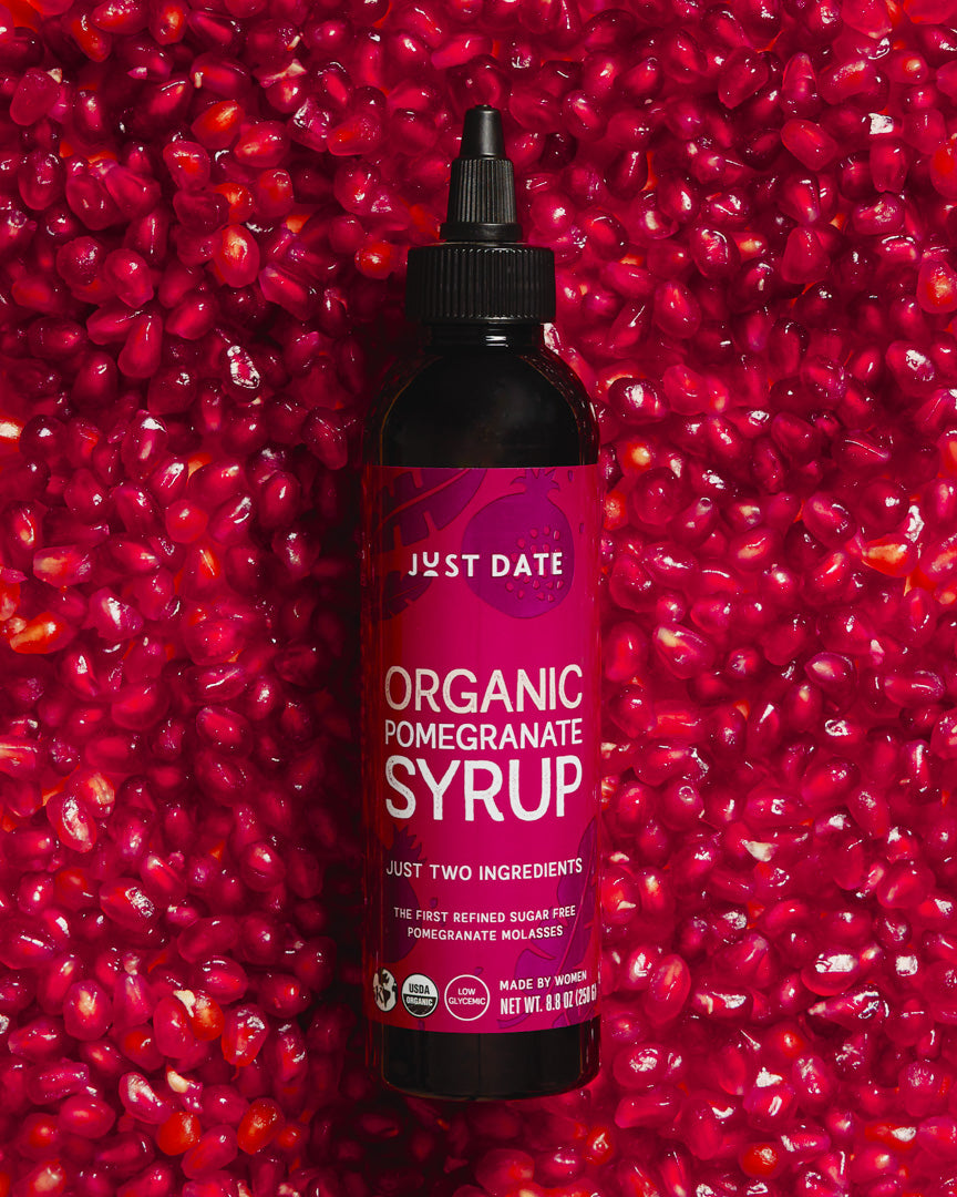 Bottle of Just Date Organic Pomegranate Syrup against a background of pomegranate seeds.