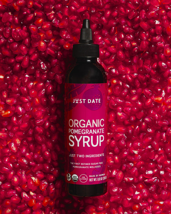 Bottle of Just Date Organic Pomegranate Syrup against a background of pomegranate seeds.