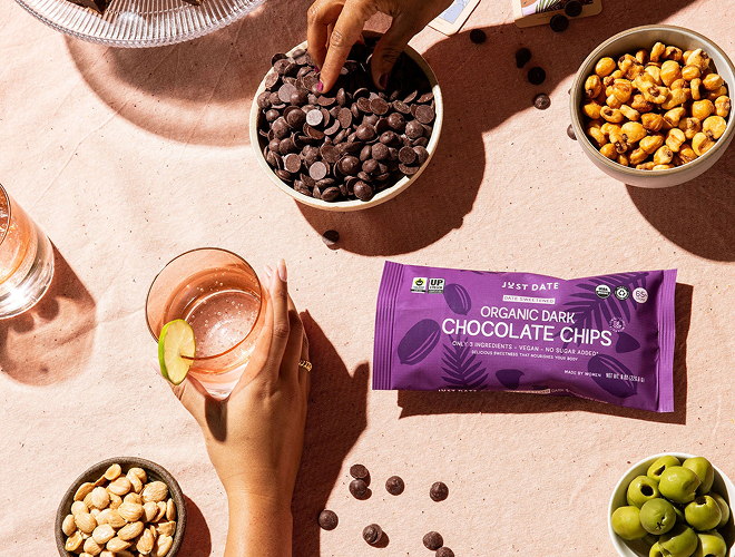 Person holding a glass of pink drink with a lime, surrounded by bowls of snacks and a package of Just Oats Organic Dark Chocolate Chips.